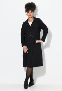 Schwarzer Trenchcoat mit doppelter Knopfleiste, tailliertem Gürtel, breiten Revers und langen Ärmeln. Trägt man über einem weißen Oberteil und kombiniert mit Knöchelstiefeln.