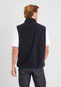 Man facing away wearing a black sleeveless fleece vest over a white short-sleeve shirt and dark jeans, against a plain gray background.