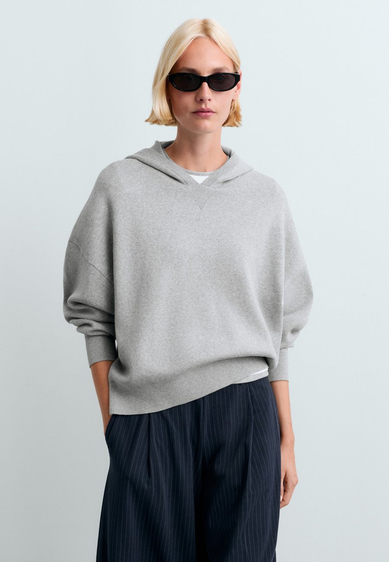 Femme blonde portant des lunettes de soleil noires, un sweat à capuche gris oversize et un pantalon large bleu marine à fines rayures, les mains dans les poches sur fond uni.