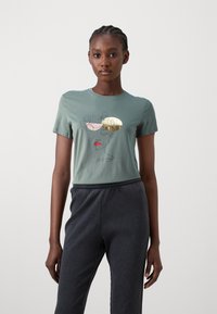 T-shirt en coton vert présentant une illustration de visage abstrait avec des accents roses, dorés et rouges. Associé à un pantalon texturé côtelé foncé.