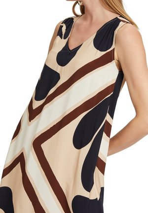 Ærmeløs kjole med V-udskæring med abstrakt geometrisk mønster i beige, brun, creme og sort båret af en kvinde med langt blondt hår.