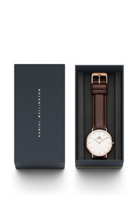 Daniel Wellington-klocka med vit urtavla, roséguld accenter och rem i brunt läder, presenterad i en mörkblå låda med mjukt inre.