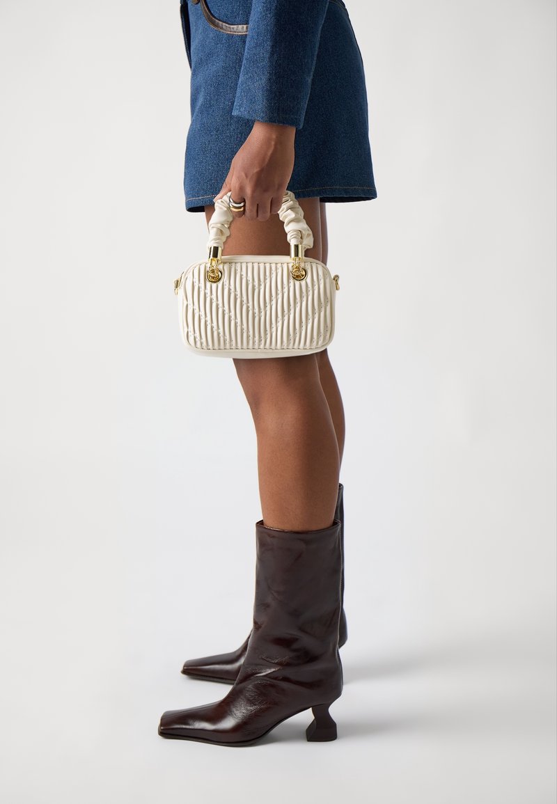 Personne portant une jupe bleue et des bottes mi-mollet marron foncé, tenant un petit sac à main matelassé crème avec des poignées froncées.