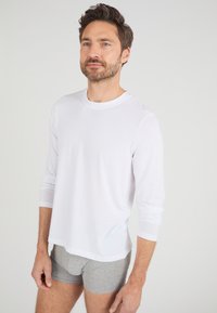 Chemise blanche à manches longues en tissu doux, col rond, coupe droite, associée à un boxer gris, se tenant contre un fond uni.