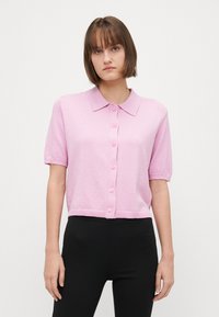 Cardigan a maniche corte di un delicato rosa chiaro, realizzato in maglia, con colletto, bottoni frontali e orlo a costine. Il tessuto ha un leggero luccichio e una morbida texture.
