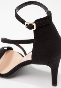 Sandale à talons hauts en suede noir avec une lanière à la cheville et un détail de boucle dorée ; présente une ouverture à l'avant et un design de talon courbé.