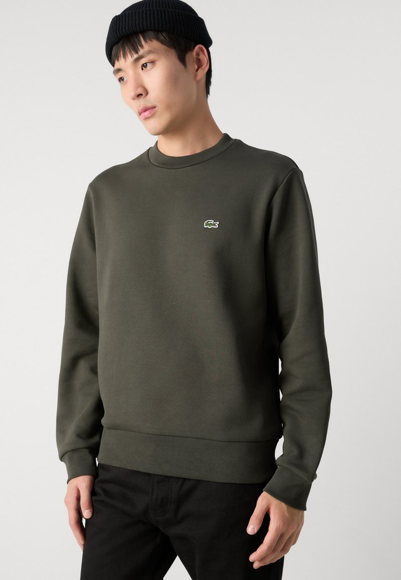 Sweatshirt vert foncé en tissu épais avec un col rond, des poignets côtelés et un petit logo Lacoste blanc sur la poitrine.