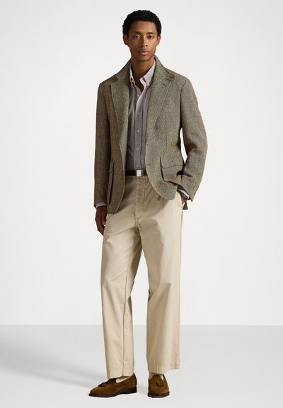 Sacou maro cu model herringbone, purtat peste o cămașă gri cu dungi, asortat cu pantaloni bej și pantofi mocasini maro. Texturi netede și croială ajustată.