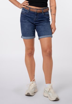 Person trägt hochgekrempelte dunkle Jeansshorts, gewebten braunen Gürtel, schwarzen Top, weiße Sneaker und zeigt Tattoos am linken Unterarm.