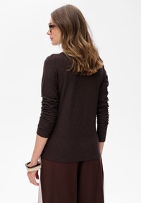 Frau mit schulterlangem braunem Haar, die Schildkrötenbrille trägt, ein strukturiertes, dunkelbraunes Langarmoberteil und dunkle Brauhosen.