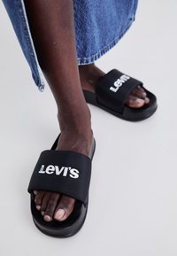 Sandales noires à glissière avec une large sangle arborant le logo "Levi's" en blanc. Texture lisse et forme ergonomique. Portées avec du denim.