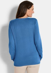 Frau mit langen braunen Haaren, die einen blauen Langarmpullover und beige Hosen trägt, leicht zur Seite gedreht und lächelnd.