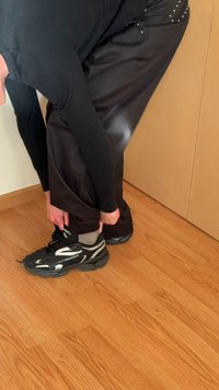 Fekete sportcipők fehér részletekkel, gumitalp, fűzős kialakítás. Egyén állítja be a fekete nadrág szárát a szürke zoknik fölött. Fapelület háttér.