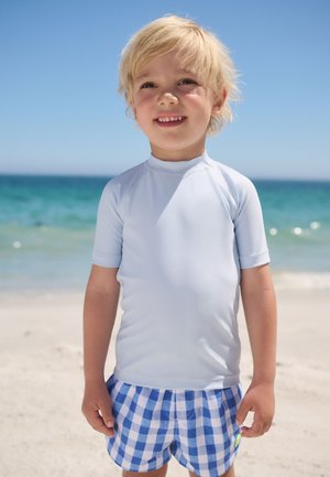Blond dziecko uśmiechające się na słonecznej plaży, ubrane w jasnoniebieską koszulkę przeciwsłoneczną i niebiesko-białe kratkowane spodenki kąpielowe, z oceanem w tle.