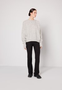 Ženska z lasmi v figi, oblečena v prevelik svetlo siv pulover, črne hlače in črne superge, stoji pred enobarvnim belim ozadjem.