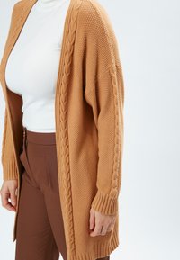 Lang, åben-front cardigan i lysebrun kabel-strik tekstur, med ribbede manchetter og søm, parret med en hvid turtleneck og brune bukser.