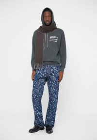 Green hoodie with a front pocket and a brown scarf. Blue denim jeans feature a white abstract pattern. Black shoes complete the outfit.