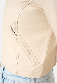 Crème-kleurige leren jas met een getailleerd ontwerp, een schuin zijrits zak en zichtbare stikdetails. G gedragen met lichtblauwe denim.
