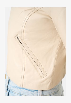 Cream-colored leather jacket featuring a fitted design, angled side zip pocket, and visible stitching details. Worn with light blue denim.