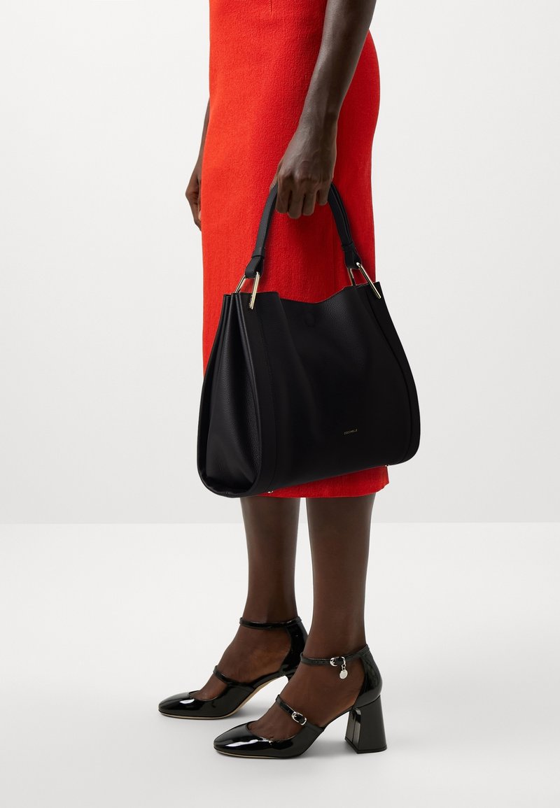 Personne en robe rouge tenant un grand sac à main en cuir noir, portant des escarpins noirs en cuir verni avec bride à la cheville et talon bloc.