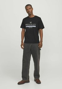 Camiseta de algodón negra con el texto "ORIGINALS" en blanco y azul, combinada con unos pantalones vaqueros anchos de color gris oscuro y botas marrones.