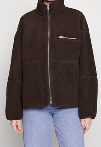 Brązowa polarowa kurtka z zamkiem błyskawicznym, wysoki kołnierz, przednia kieszeń na zamek oraz dwie duże kieszenie z przodu. Luźny krój i teksturowany, miękki materiał.