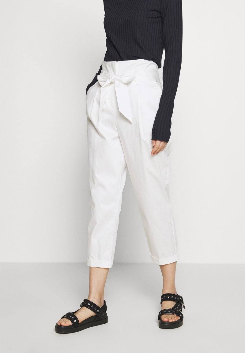 Witte, hooggetailleerde broek met een strikdetail aan de voorkant, verkorte lengte en een gladde textuur. Gecombineerd met zwarte sandalen met hardware-accenten.