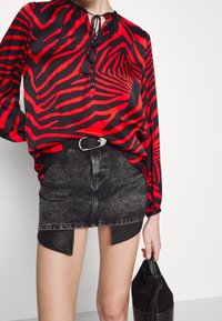 Femme portant un chemisier rayé zèbre rouge et noir, une jupe en jean noire délavée à l'acide avec ceinture, tenant un sac à main en cuir noir.