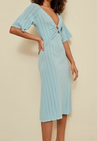 Robe tricotée bleu clair avec un décolleté en V profond, des manches courtes et flottantes, et un détail de nœud à l'avant. Présente un motif texturé en zigzag et un ourlet festonné.