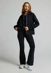 Zwarte waterdichte jas met een ritssluiting aan de voorkant, capuchon en logo. Gedragen over een zwart top en uitlopende broek, gecombineerd met zilveren en zwarte sneakers.