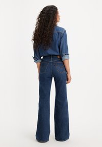 Žena s dlhými kudrnatými vlasmi, oblečená v modrej riflovej košeli a modrých džínsach s vysokým pásom a širokými nohavicami, stojí s chrbtom otočeným dopredu.