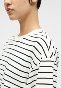 Νεαρό άτομο με κοντά μαλλιά, που φοράει λευκό τοπ με ομοιόμορφες μαύρες οριζόντιες ρίγες, εμφανιζόμενο από το πλάι και κομμένο στο ύψος του λαιμού.