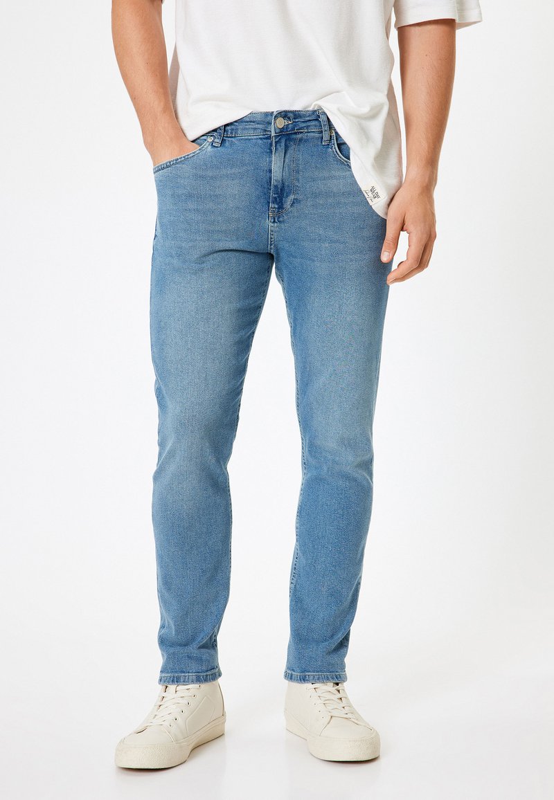 Jeans in denim azzurro chiaro con vestibilità slim, cinque tasche, passanti per la cintura e chiusura a bottone. Indossati con sneakers bianche e una camicia bianca dalle linee morbide.