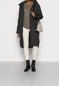 Manteau long matelassé noir avec capuche, motif piqué. Pull vert olive et pantalon skinny crème. Bottines noires à talons, portant un petit sac gris.