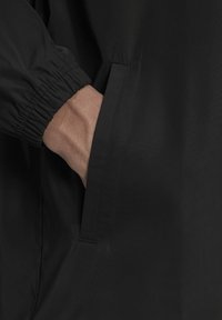 Svart jacka med strukturerad tyg, elastiska ärmslut och en sidoficka integrerad i designen. Närbild på ärm och ficka.