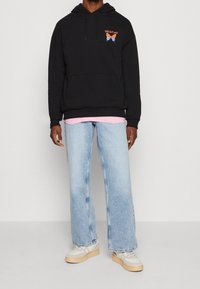 Felpa nera in cotone con tasca frontale e design grafico; indossata sopra una maglietta rosa e abbinata a jeans a zampa d'elefante azzurri chiari e sneakers.