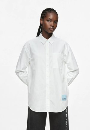 Femme noire aux cheveux tressés portant une chemise blanche oversize boutonnée avec une poche avant et un pantalon noir avec texte blanc sur le côté.