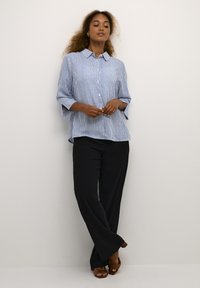 Camicia a righe blu e bianche con colletto, vestibilità ampia e maniche a tre quarti, abbinata a pantaloni neri a gamba larga e tacchi marroni.