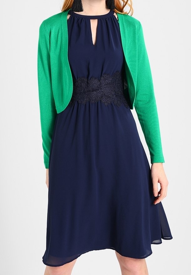 Femme portant une robe sans manches bleu marine avec un détail en dentelle à la taille et un cardigan court vert vif, debout sur un fond uni.