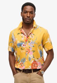 Chemise à manches courtes, boutonnée, de couleur jaune vif avec un motif floral comportant des fleurs roses, rouges et bleues. Tissu léger, coupe décontractée.