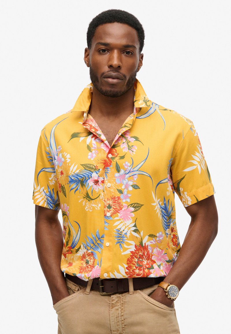 Korte mouwen, button-up shirt in felgeel met een bloemenpatroon van roze, rode en blauwe bloemen. Lichtgewicht stof, ontspannen pasvorm.