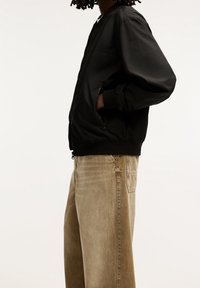 Svart bomberjacka med en slät textur och dragkedja framtill, parat med ljusbruna vida jeans som har en bakficka.