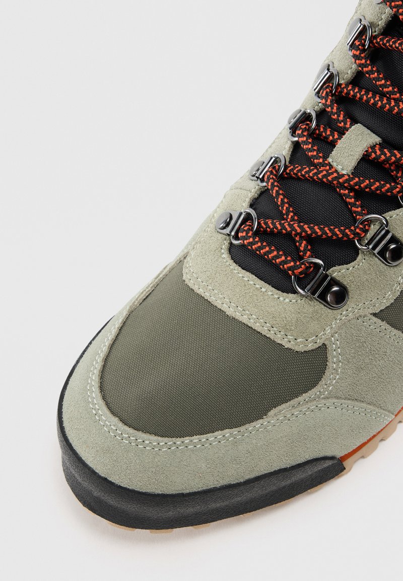 Grüne Wildleder- und Stoff-Wanderschuhe mit orangefarbenen Schnürsenkeln, Metallösen und einer strukturierten Gummisohle. Auffälliges Design für den Outdoor-Einsatz.