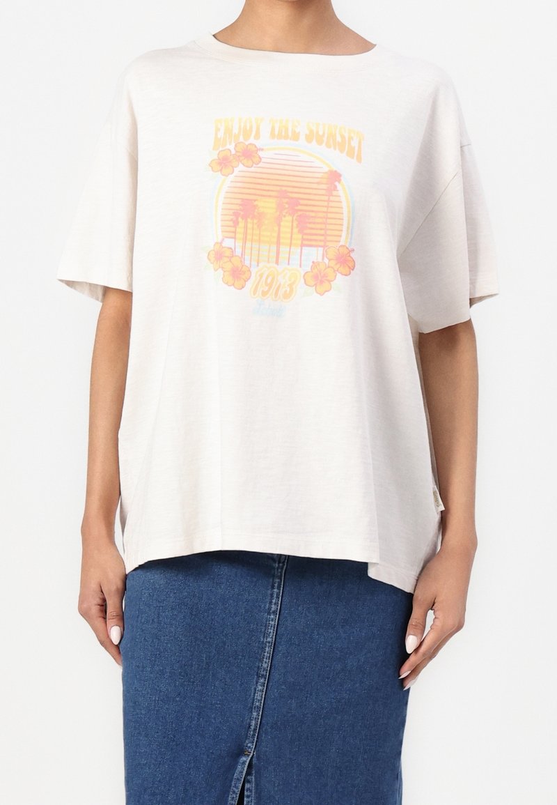 T-shirt blanc oversize avec coucher de soleil jaune et orange, palmiers, fleurs d'hibiscus, texte "Enjoy the Sunset 1973", porté avec une jupe en jean bleue.