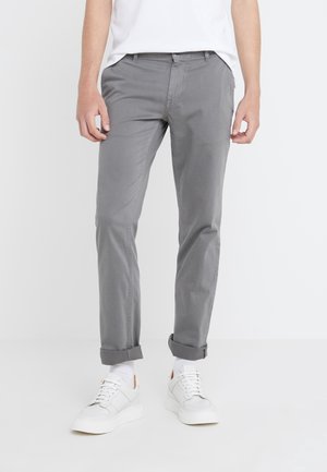 Mand iført opsmøgede grå bukser, hvide sneakers og hvide sokker, stående mod en ensfarvet hvid baggrund.