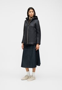 Νεαρή γυναίκα που φοράει μαύρο καπιτονέ μπουφάν με κουκούλα, σκούρο denim φούστα μέσης μήκους, λευκές κάλτσες και μπεζ αθλητικά παπούτσια, όρθια και κοιτάζει πλάγια.