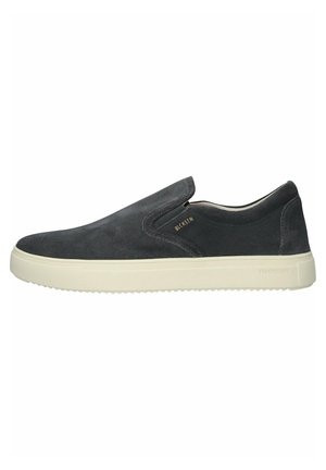 Zapato negro de ante sin cordones con acentos elásticos en los laterales, suela de goma blanca y una marca mínima. Textura suave y diseño estilizado.