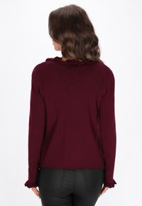 Bordowy prążkowany top z długim rękawem, z falbanami przy dekolcie i mankietach; ma luźny krój i teksturowany materiał z lekko skróconym dołem.