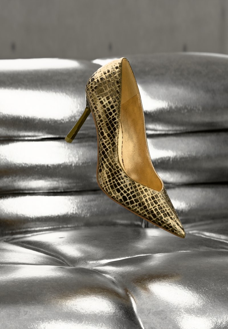 Chaussure à talon haut dorée à bout pointu, fabriquée en peau reptile synthétique texturée, avec un talon aiguille élégant, posée sur une surface argentée brillante.