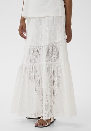 Osoba ubrana w białą, koronkową spódnicę maxi z geometrycznym wzorem, zestawioną z białymi sandałami ozdobionymi srebrnym akcentem na pasku.